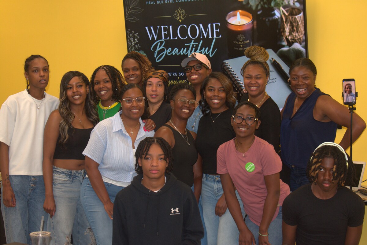 Group portrait of the women and young people who attended the first Heal Blk Gyrl gathering, smiling together in front of a Welcome Beautiful screen
